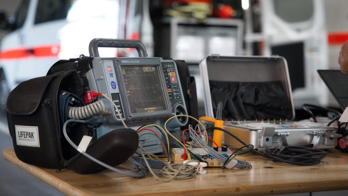 Defibrillator mit angeschlossenen Prüfleitungen auf dem Prüftisch, daneben der geöffnete Koffer mit der Prüftechnik