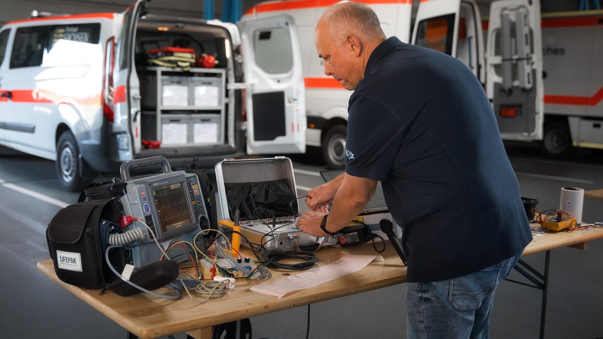 Ralf Gäbler prüft einen Defibrillator am Prüftisch, angeschlossen an ein Sicherheitsprüfgerät, im Hintergrund Rettungswagen