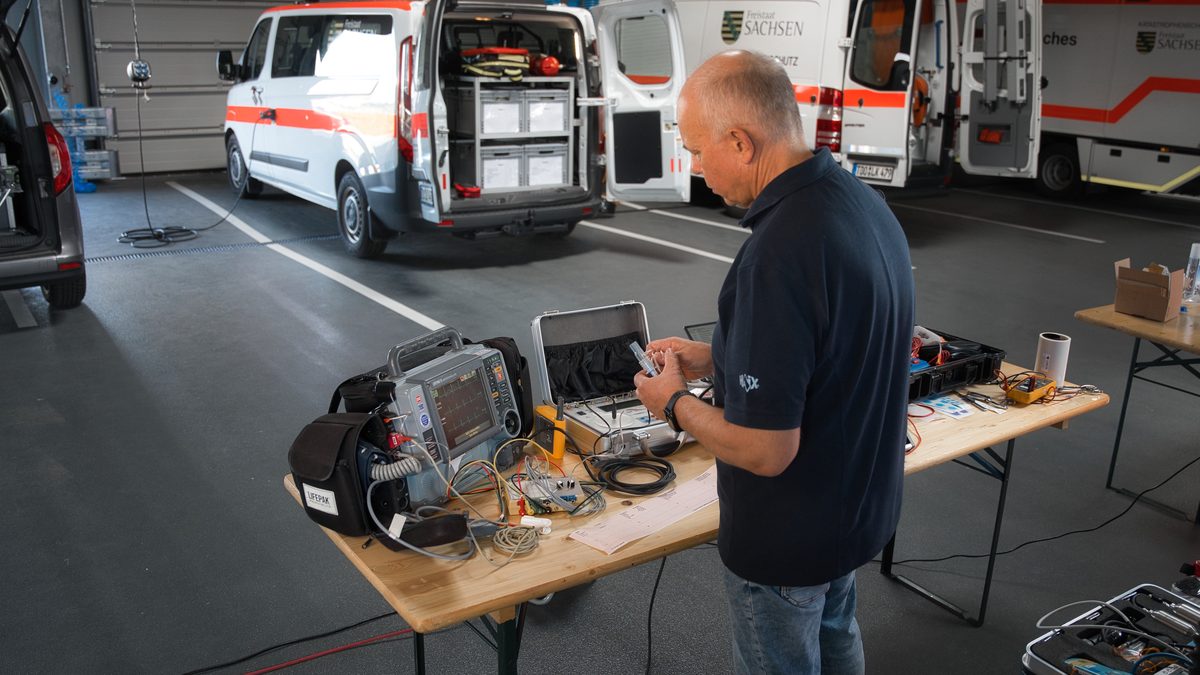 Prüfplatz in der Fahrzeughalle: Ralf Gäbler am Prüftisch mit Defibrillator und Messtechnik, dahinter zwei Rettungswagen